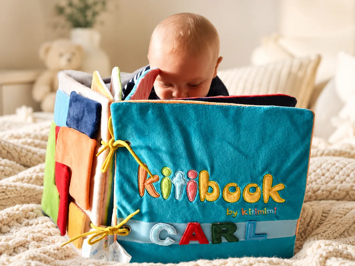 Cadeau de naissance personnalisé : jeune bébé découvrant le livre en tissu Kitibook au prénom appliqué, sur un plaid crème